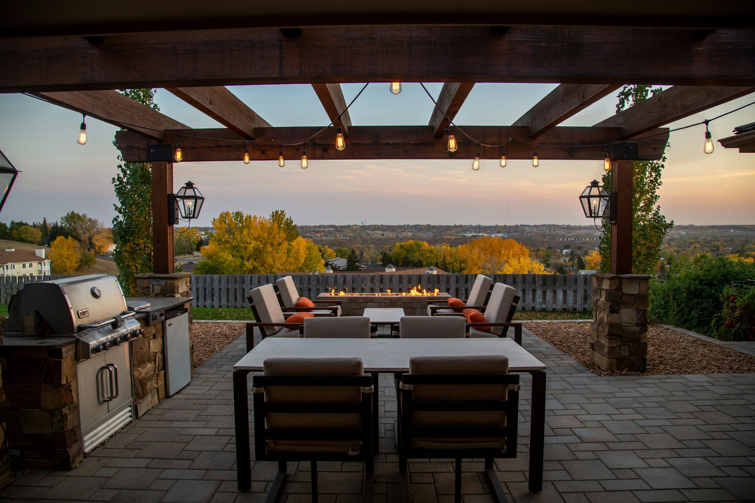 Back patio with an outdoor kitchen, grill, pergola, hanging lights, and 2 sets of outdoor dining tables with chairs.